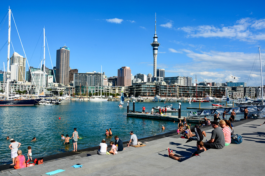 View from the Wynyard Quarter in Auckland, with people, residential and commercial buildings, Sky Tower and boats.