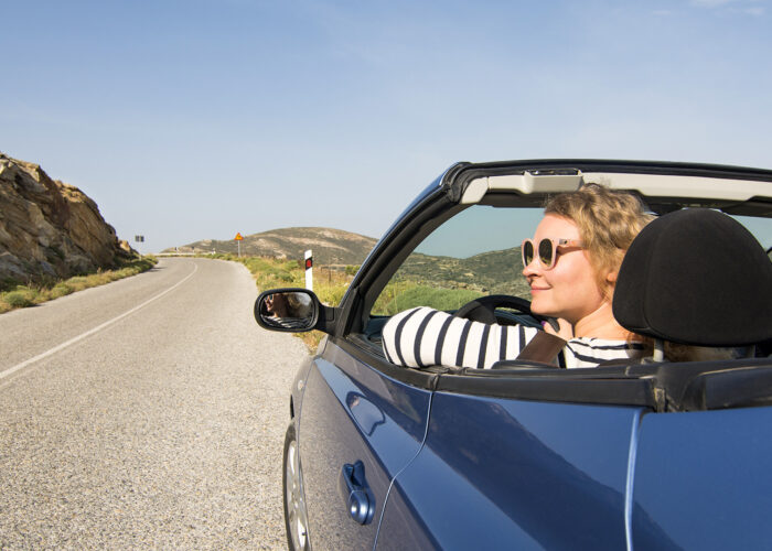 Young blonde woman driving in convertible blue car without roof on mountain road in Naxos island, Greece.