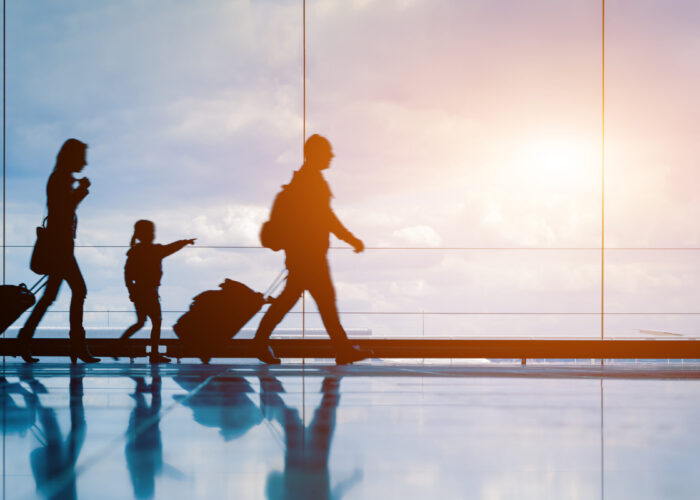 Silhouette of family walking along a glass-walled walkway in airport, rolling their suitcases