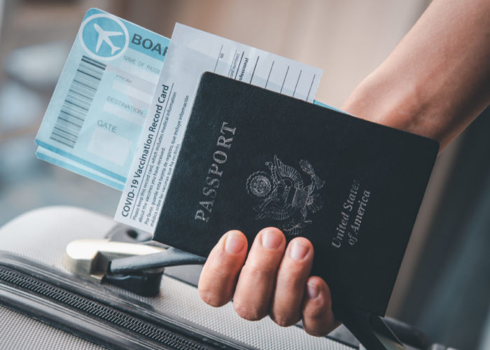 A person holding their COVID-19 vaccine card, passport, boarding pass, and suitcase