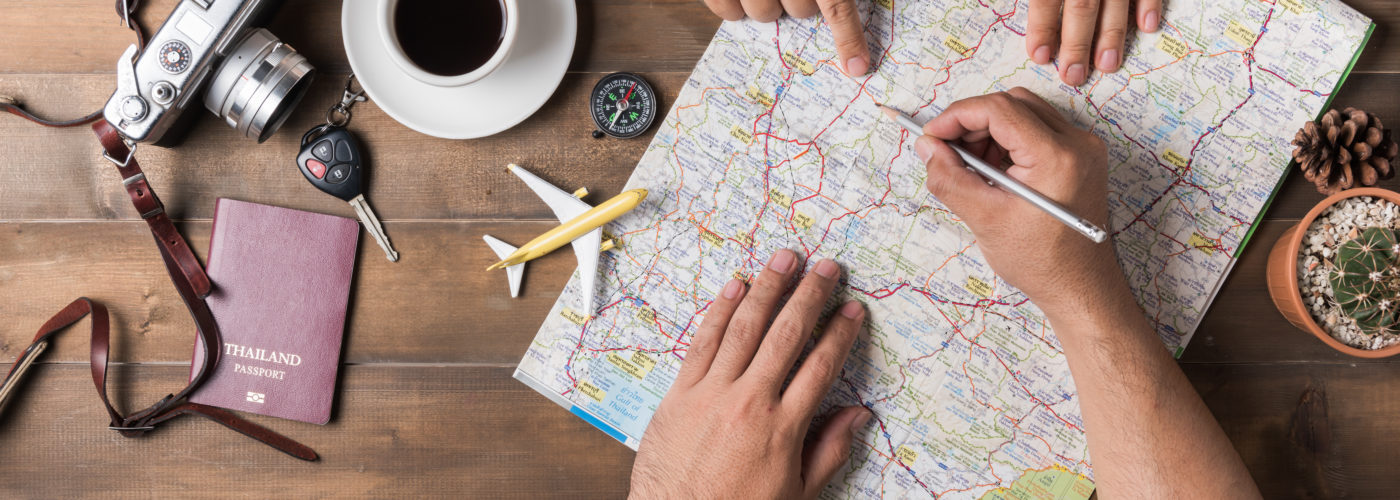 Overhead view of two people planning a vacation on a map surrounded by a cup of coffee, a model plane, a camera, and a passport cover