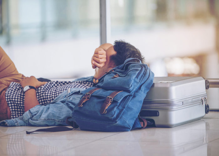 Person laying on the airport floor, resting their head on a backpack and a suitcase
