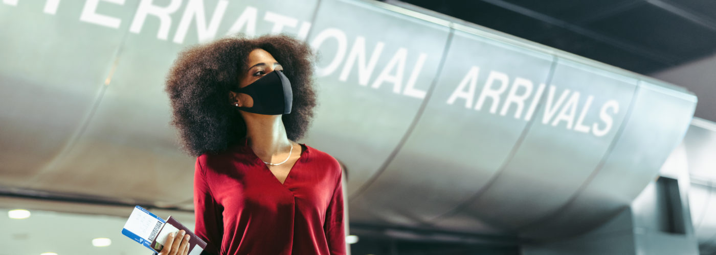 Woman standing at international arrivals terminal holding a boarding pass and wearing a face mask