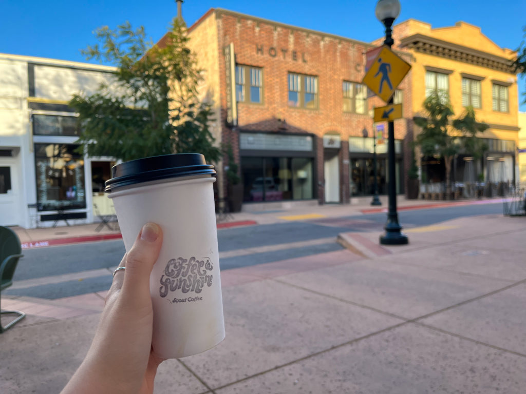 Hand holding a cup of coffee with the Scout Coffee logo in front of a sunny street