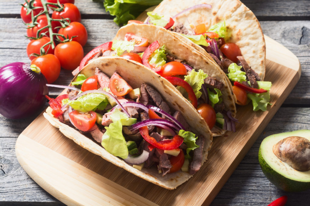 Three pork tacos on a wooden cutting board surrounded by fresh ingredients