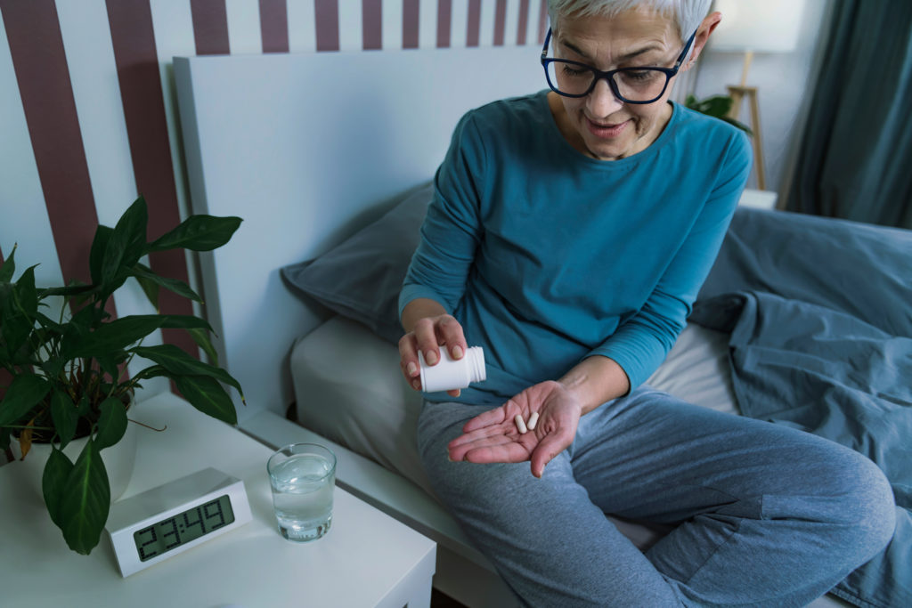 Older woman sitting on a bed and pouring melatonin pills from a bottle into her hand