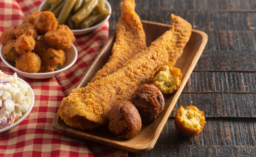 Plate of fried catfish and hush puppies next to smaller plates of hush puppies, green beans, and coleslaw on a wooden table and a red and white plaid table cloth 