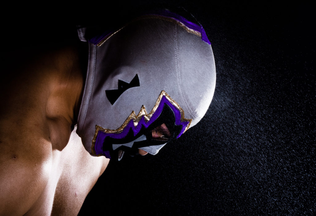 Luchador in a white and purple mask in a dark room
