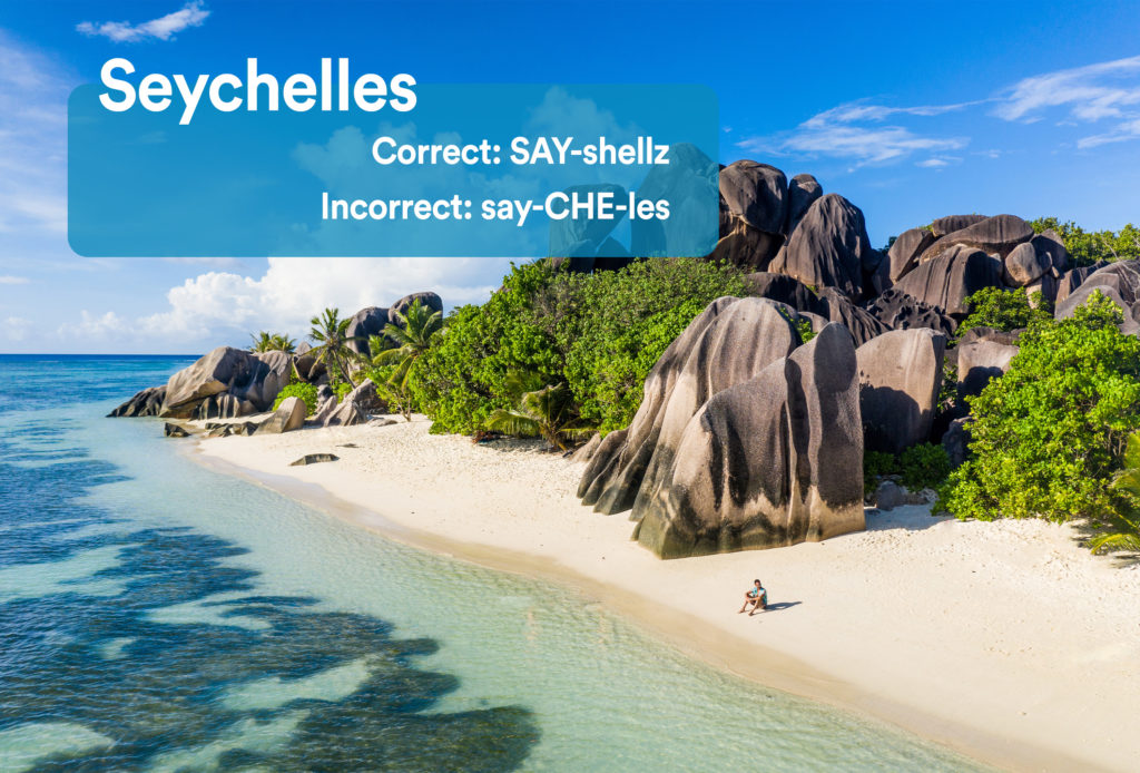 Person sitting on the beach near the water and large rock formations on an island in the Seychelles with graphic overlay showing the correct and incorrect way to pronounce "Seychelles"
