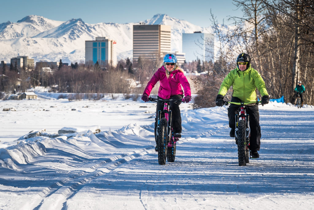Two people using fat tire bikes on a biking trail with the Anchorage, Alaska skyline in the background