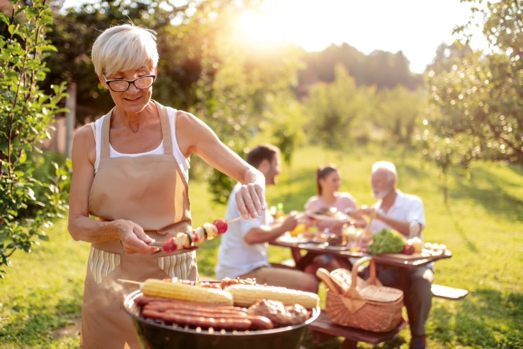 Older woman grilling meat at a circular grill with friends at table in background