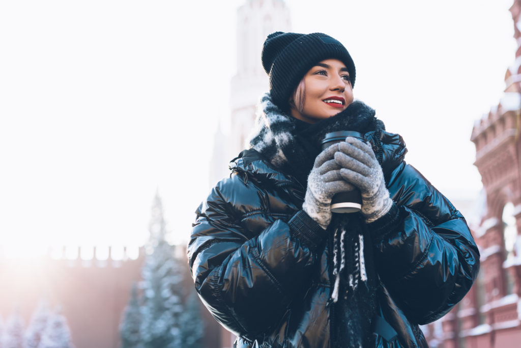 Woman wearing a winter jacket, hat, scarf, and gloves holding a travel mug of a hot beverage in a winter city scene