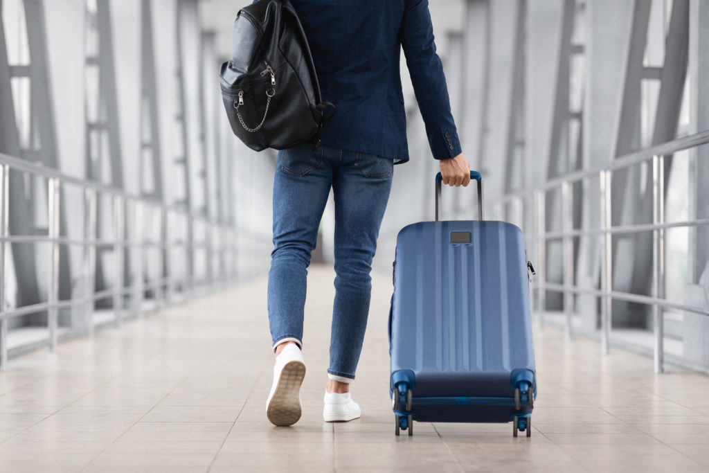 Man pulling blue suitcase down a glass hallway in an airport