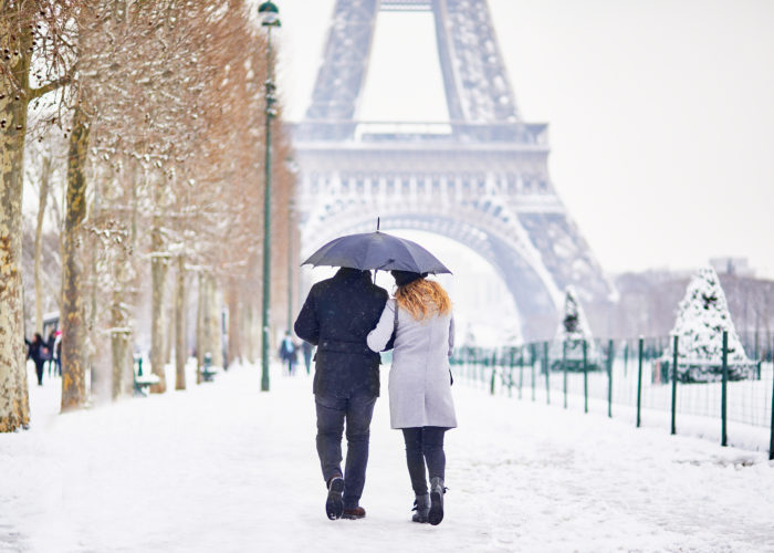 Couple walking along a tree-lined path toward the Eiffel Tower in the snow