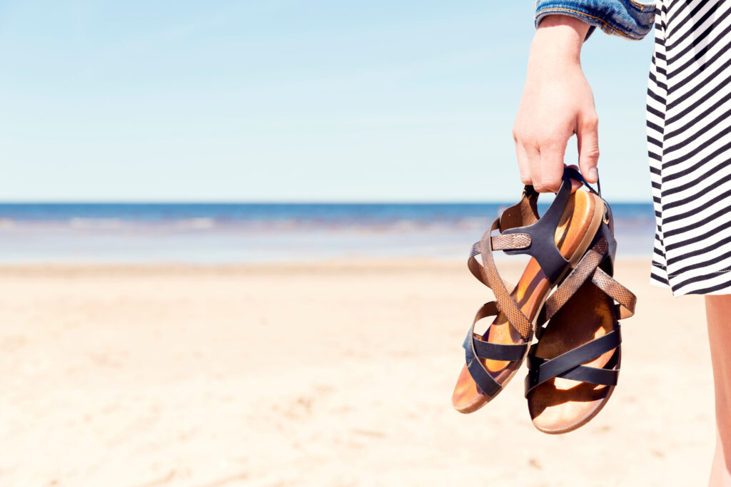 Close up of person holding their strappy sandals in their hand on the beach