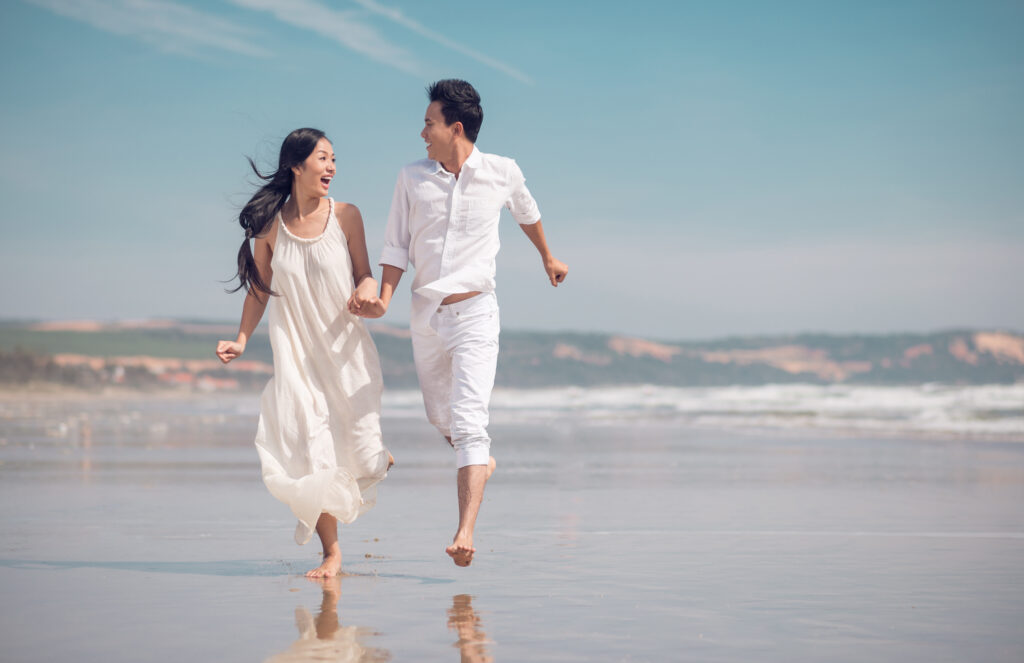 Man and women holding hands and running on the beach, wearing coordinating white linen outfits
