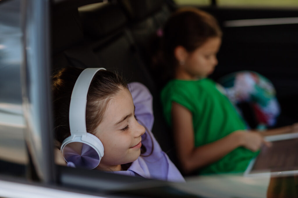 Two children in the backseat of a car as seen through the car window, one listening to music on headphones and the other on a tablet device