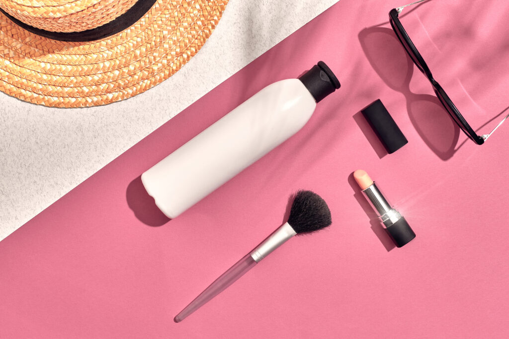 Makeup, sunscreen, sunglasses, and a sunhat laid flat on a pink background
