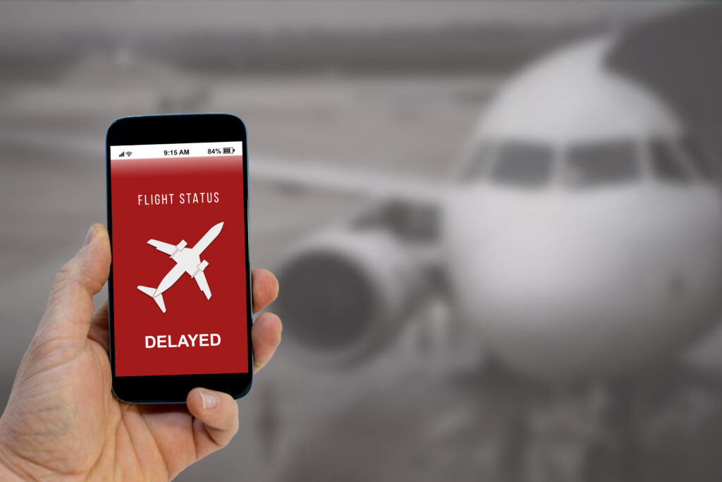 Close up of person holding phone with red message reading "Flight Status: Delayed" with an out of focus airplane in the background