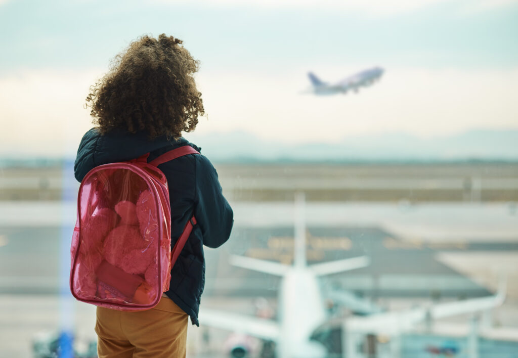 Child wearing pink backpack and looking out window at airport at plane taking off