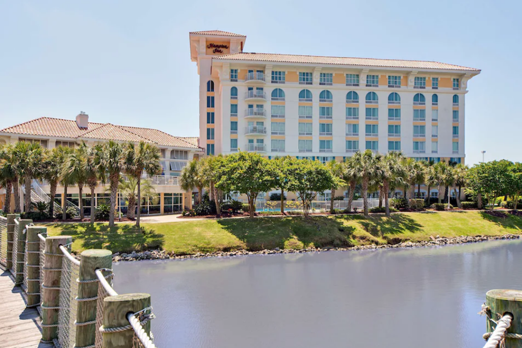 Exterior of the Hampton Inn Myrtle Beach Broadway at the Beach with water in the foreground and palm trees in front of the building