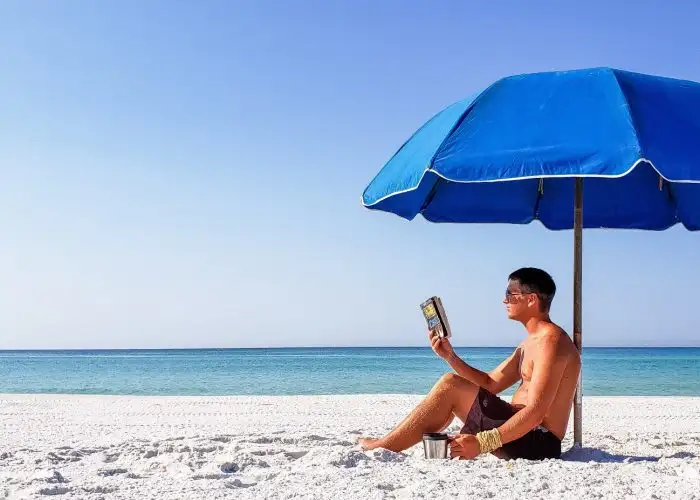Man reading a paperback book under a beach umbrella.
