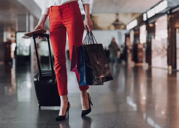 Woman, seen from the waist down, wearing high heels and carrying a rolling suitcase and shopping bags in an airport