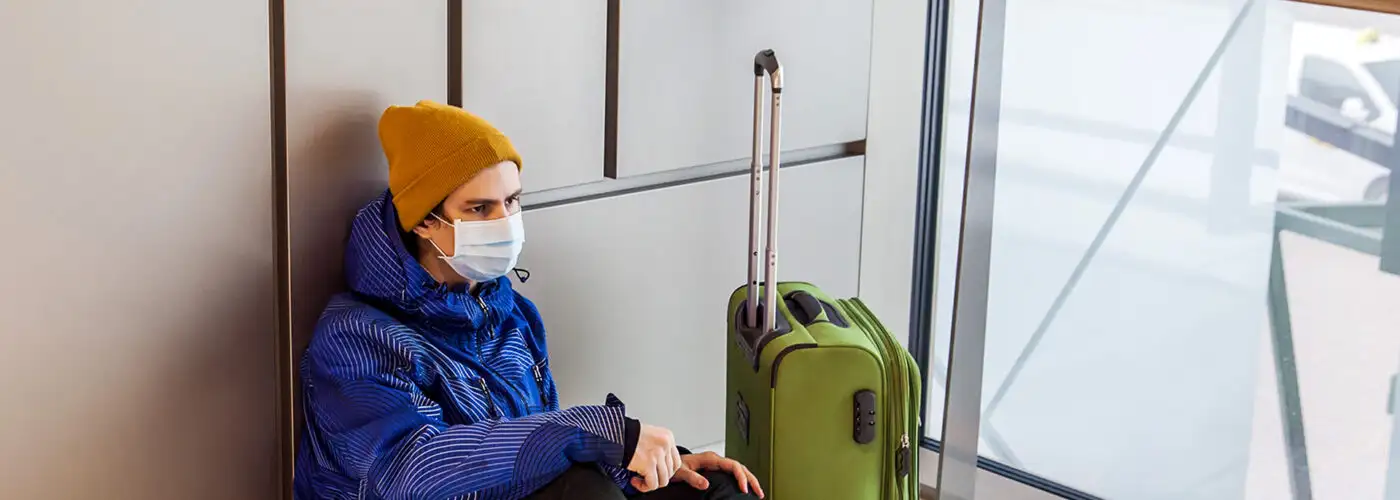 boy with a green suitcase is sitting on floor in waiting area wearing protection mask