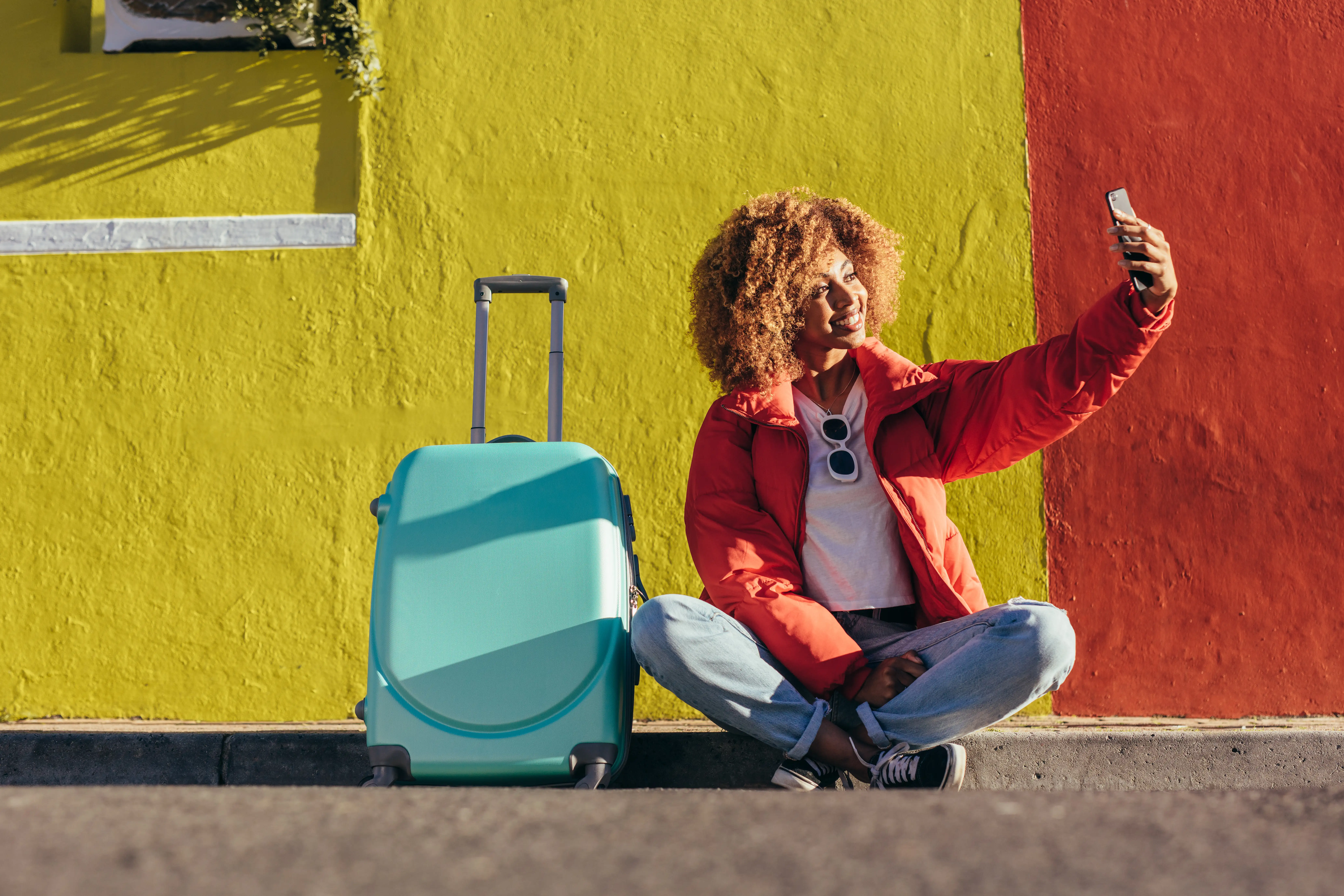 Woman taking selfie in front of a brightly colored wall next to a suitcase