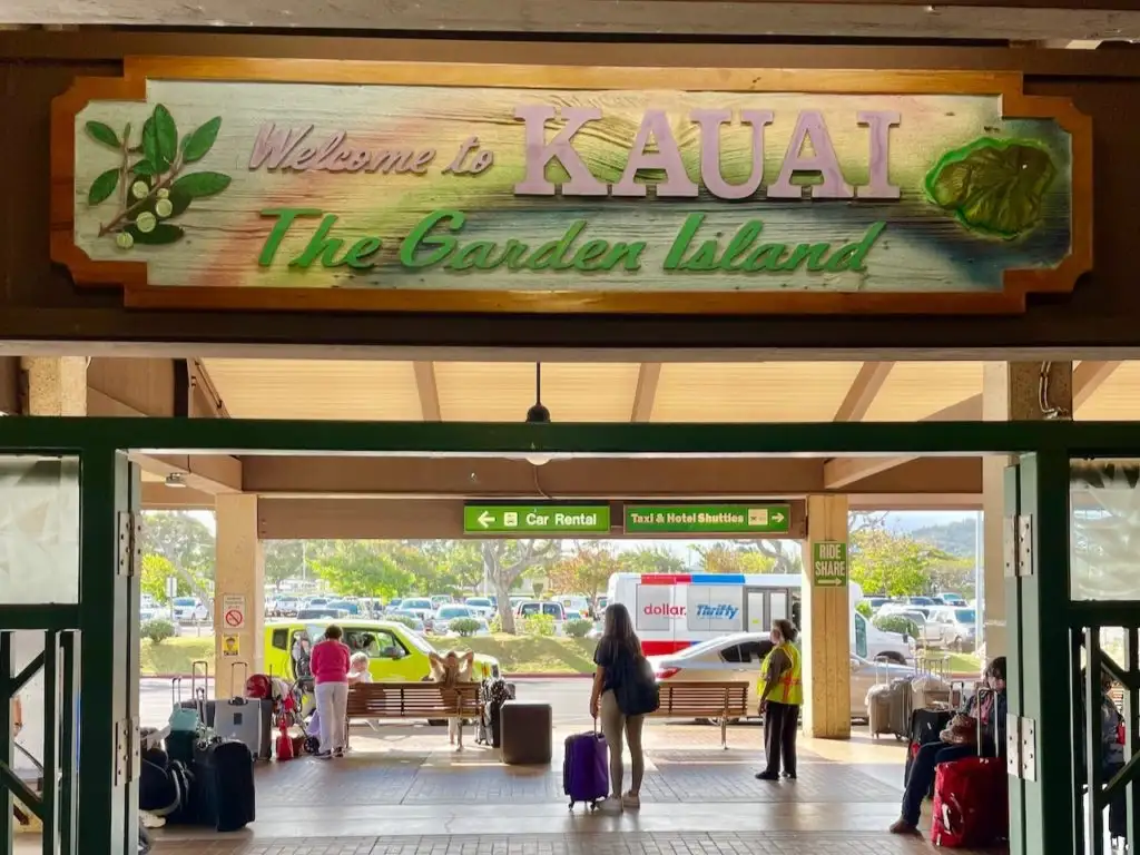 Tourists with suitcases waiting beneath a "Welcome to Kauai" sign