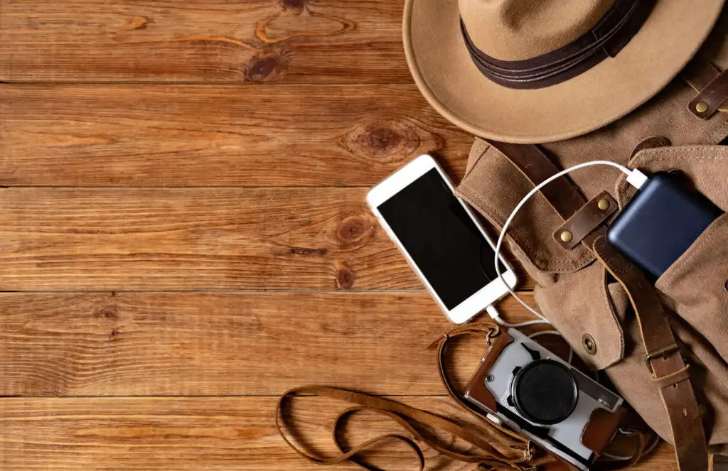 A backpack on a wooden background with a phone, power pack charger, a camera, and a hat spilling out of it
