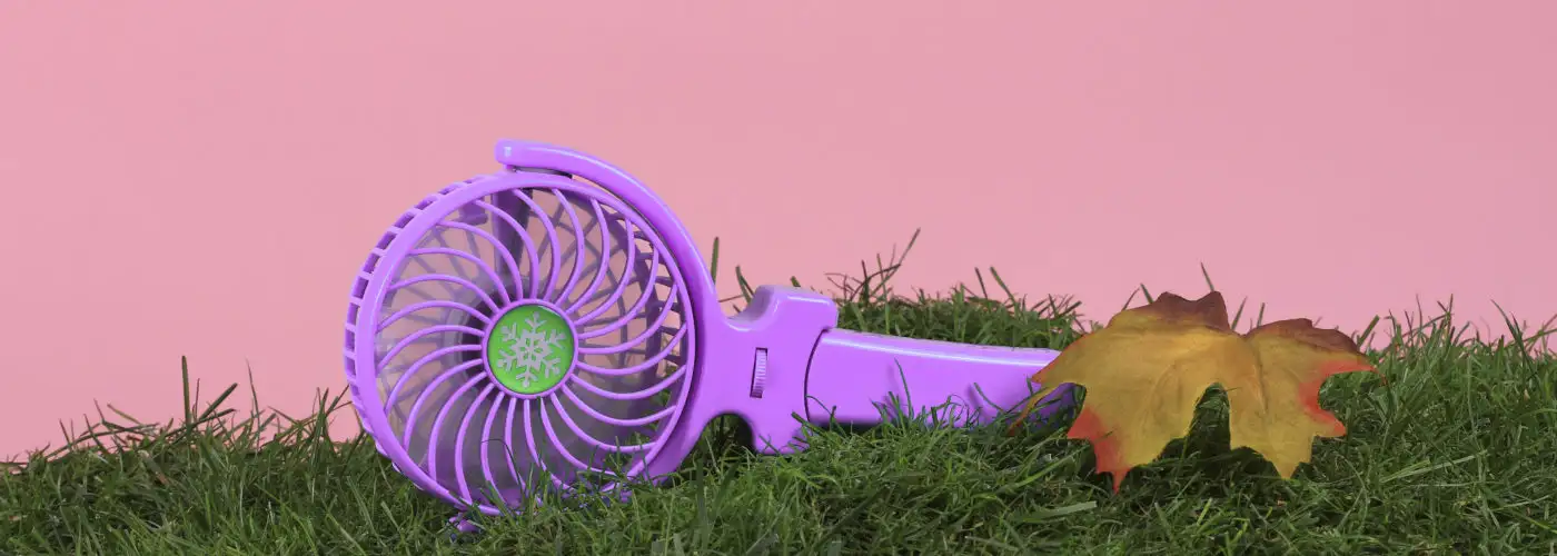 Handheld electric fan on a bed of grass with a pink backdrop