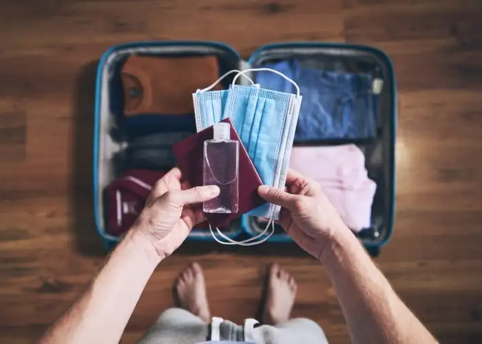 Person holding a face mask, passport book, and hand sanitizer over a packed suitcase on the floor