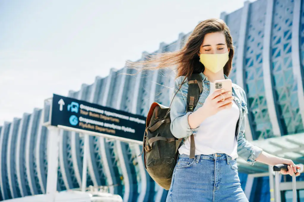 Woman walking away from an airport terminal, wearing a face mask and using her smartphone