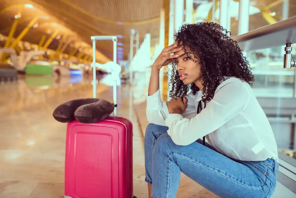 Frustrated woman sitting on floor of airport next to red luggage and neck pillow