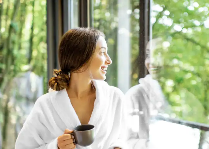 Woman holding a coffee mug looking out a large window while wearing a white bathrobe