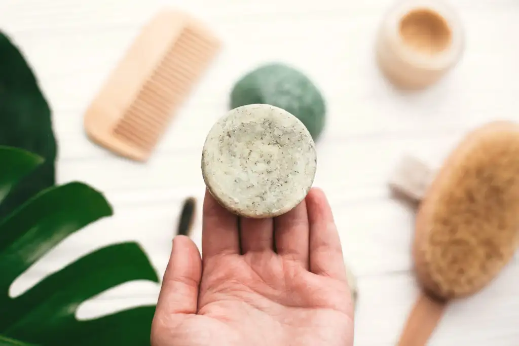 Close up of person's hand holding a green circular shampoo bar with other wooden bathing products out of focus in the background