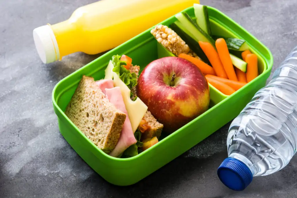 Fruit, vegetables, and a sandwich packed in a reusable plastic lunchbox next to a water bottle and a bottle of orange juice 