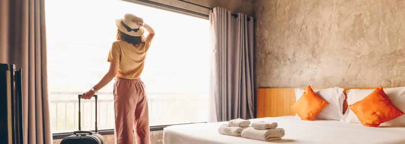Woman standing in a hotel room and looking out the window, holding the handle of her luggage