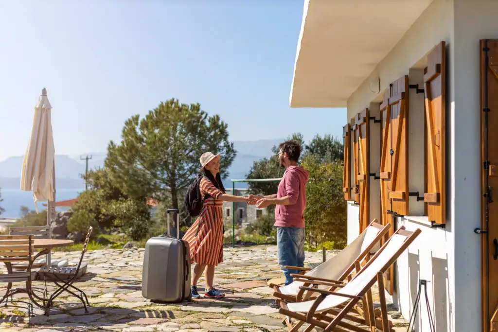 Two people shaking hands, standing in front of a vacation rental property in the mountains and next to a grey rolling suitcase
