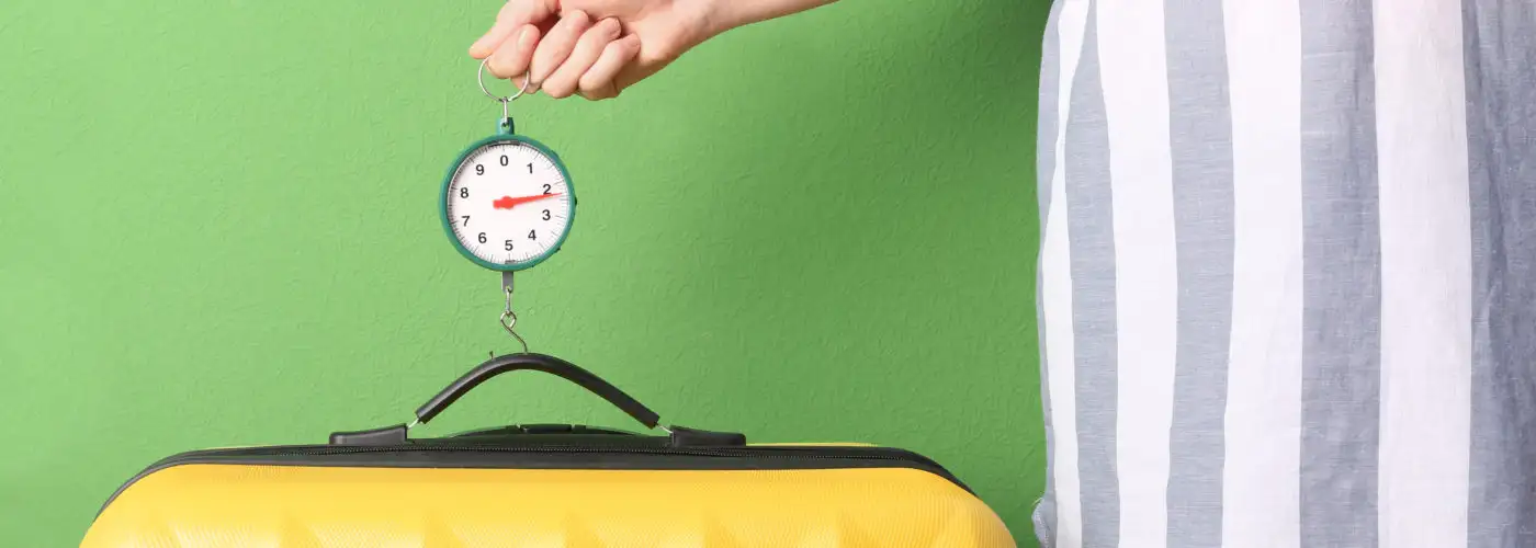 Close up of person wearing a dress and using a luggage scale to weigh a yellow suitcase against a bright green wall