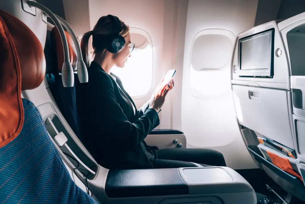 Woman sitting in window seat on airplane listening to media on her phone using noise cancelling headphones