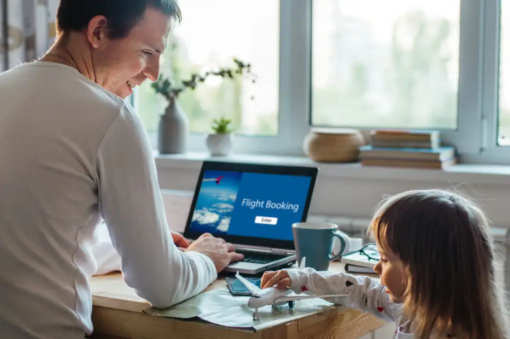 Father booking flights on a laptop while daughter plays with model plane on his desk