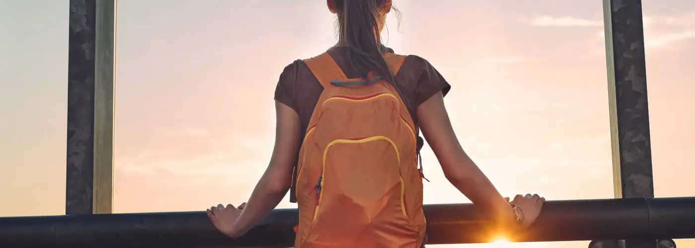 Woman standing with backpack looking out a window at sunset in an airprot
