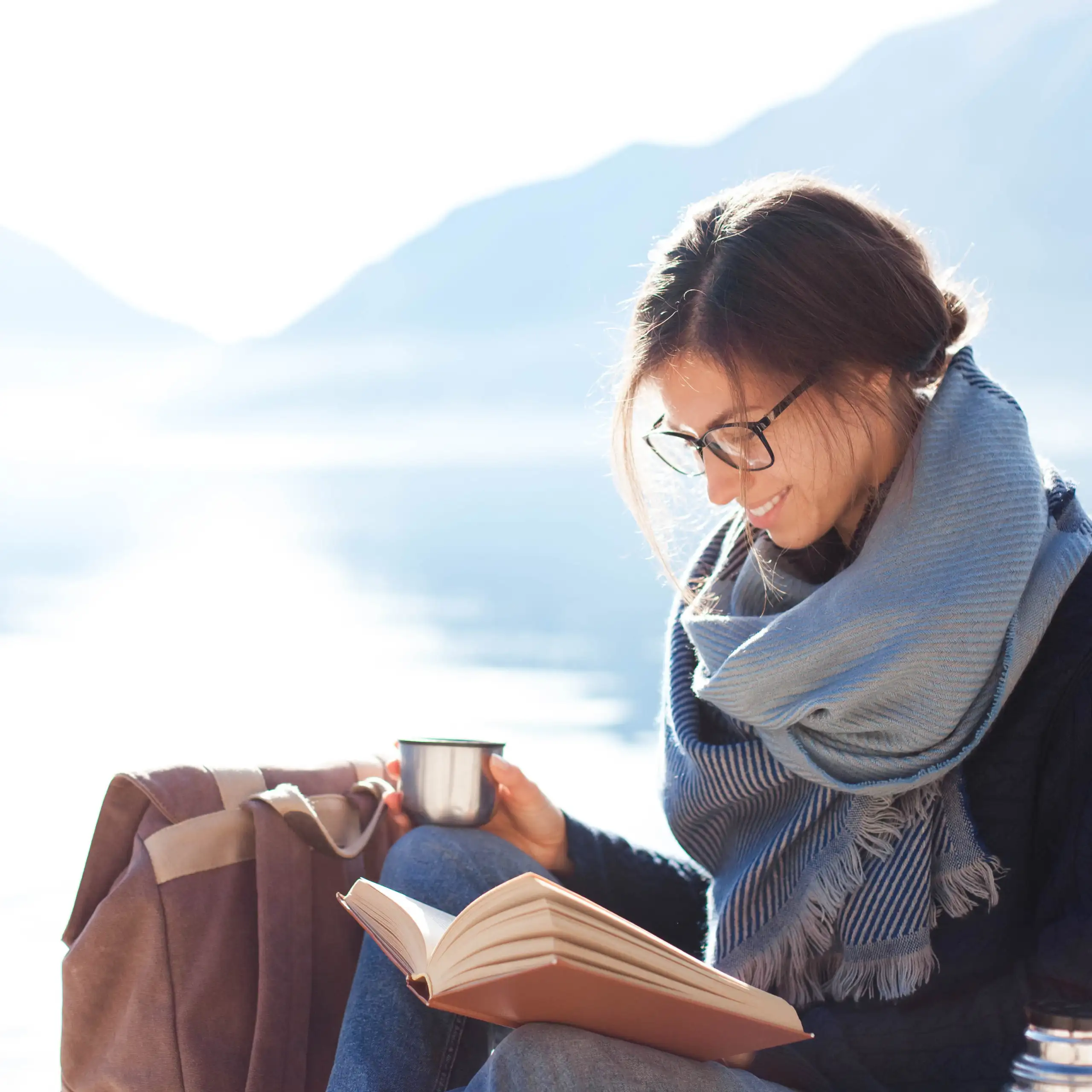 Woman wearing a blanket scarf, drinking from a mug, and reading a journal next to a lake