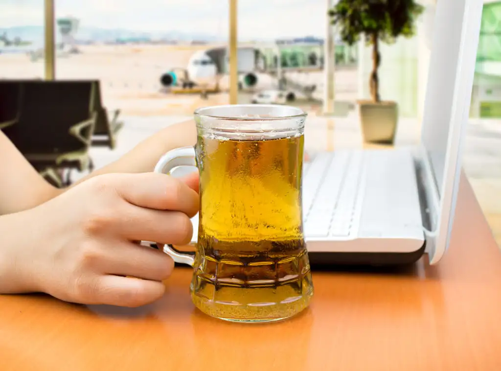 Close up of person drinking a large stein of beer while working on a laptop at an airport bar