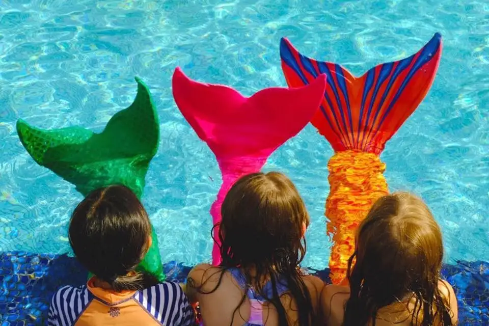Three children wearing mermaid tails sitting on the edge of a pool