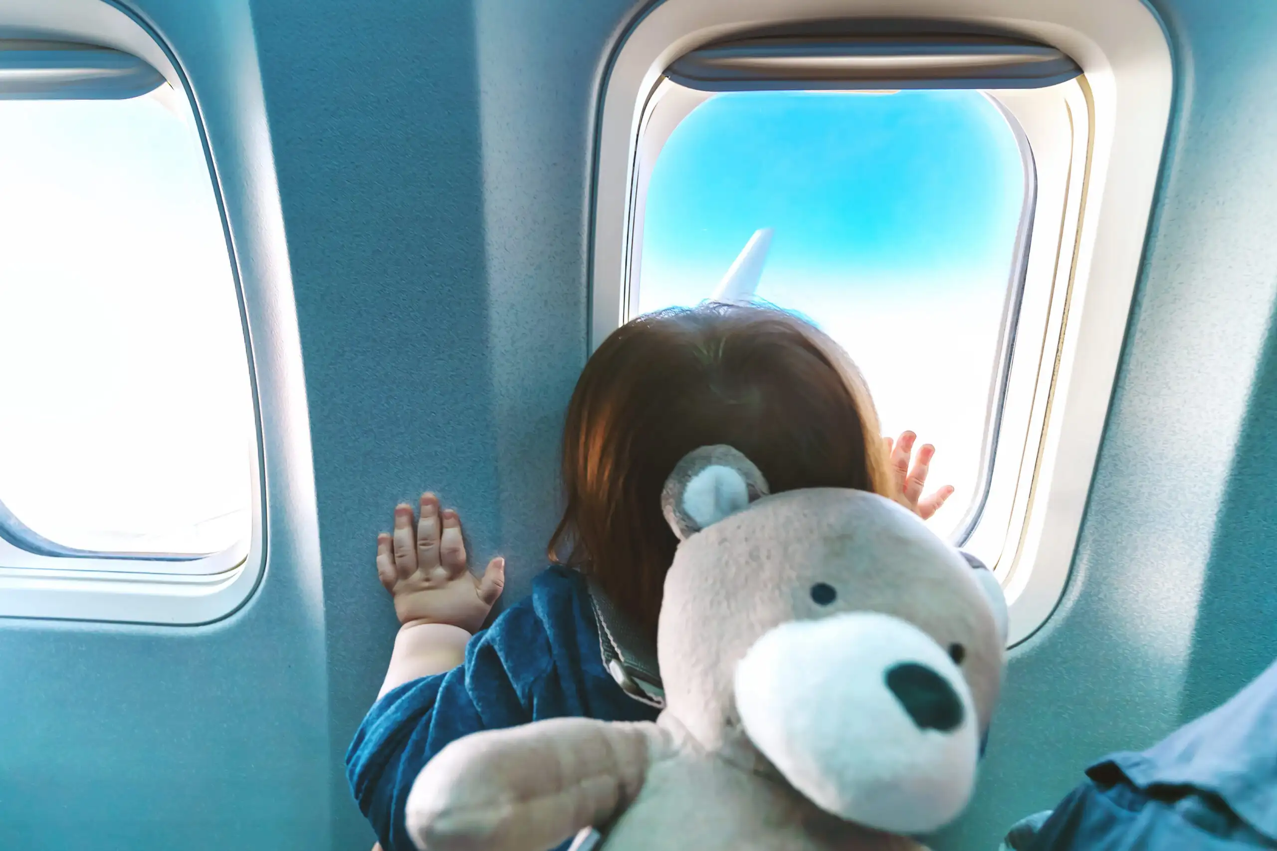 Toddler looking out airplane window with teddy bear