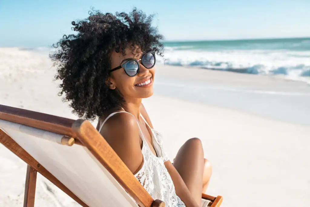 Young woman sitting on the beach in a beach chair, wearing sunglasses and smiling at the camera