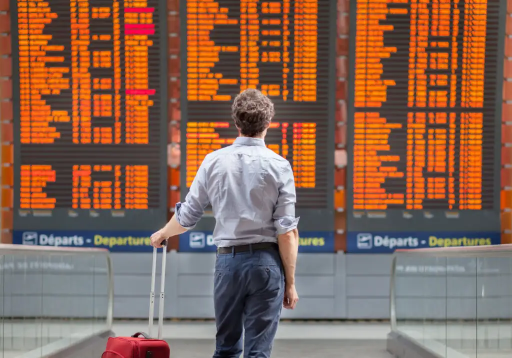 Man waiting with rolling luggage, looking up at departure board at an airport